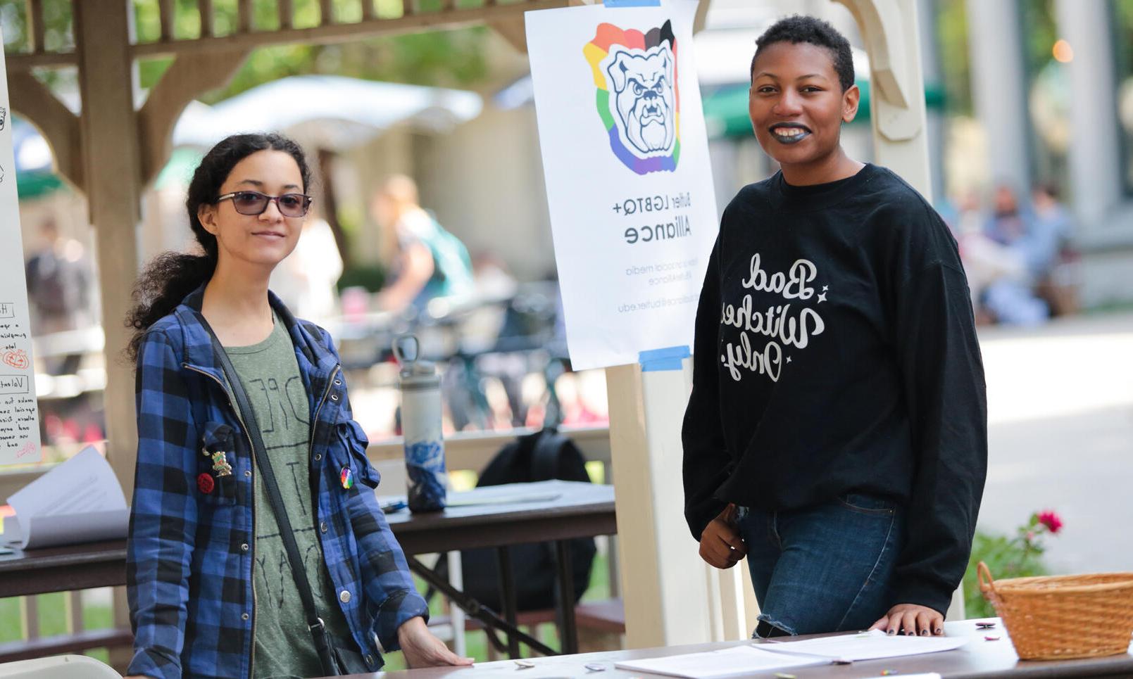 两个LGBTQ+联盟的成员拿着那个牌子, 一个穿着黑色运动衫，上面写着“只允许坏女巫”, 另一个戴着墨镜，穿着蓝黑相间的格子衬衫
